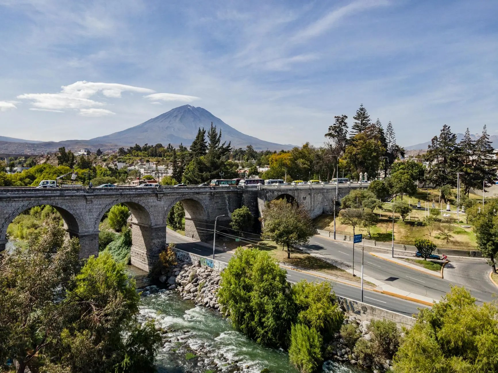 A scenic view of a bridge arching over a river, with lush greenery lining the banks and a tall volcano in the background under a clear blue sky.