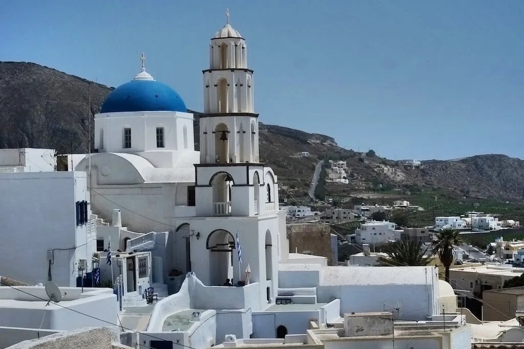 A scenic view of traditional white-washed buildings and blue-domed churches in a Santorini village, with a mountainous backdrop and clear blue sky.