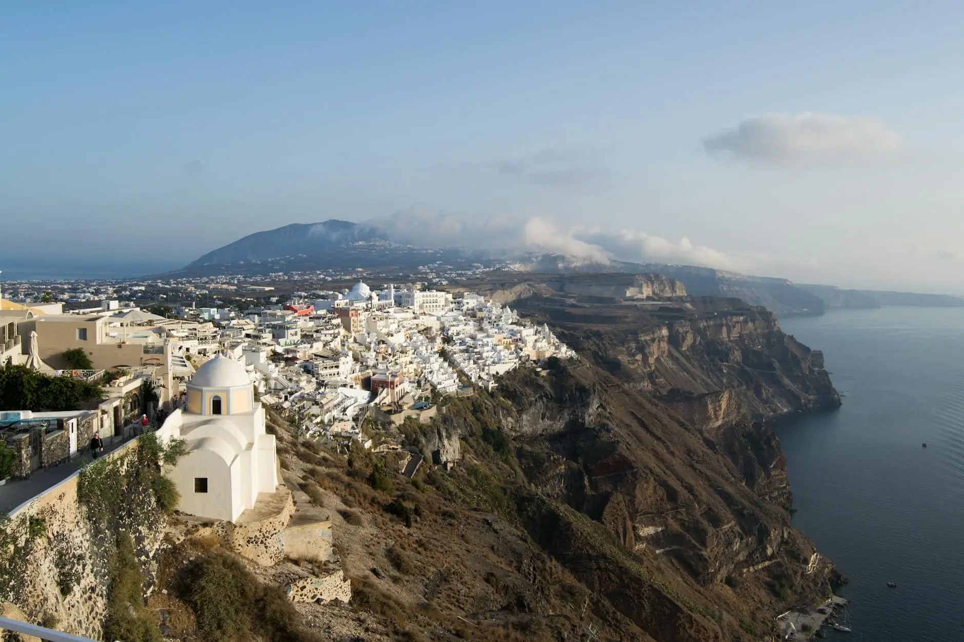A panoramic view of Santorini featuring whitewashed buildings on a cliffside overlooking the Aegean Sea, with a mountainous backdrop under a clear sky.