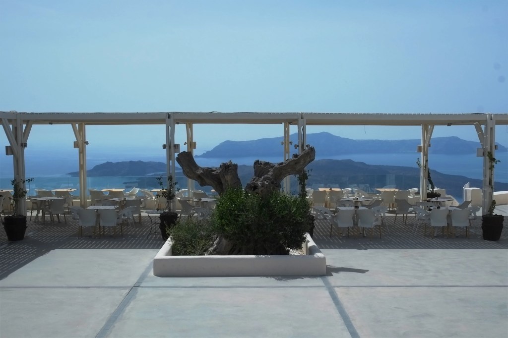 A scenic view of a restaurant terrace in Santorini, featuring white furniture, plants, and a magnificent backdrop of the Aegean Sea and distant islands under a clear blue sky.