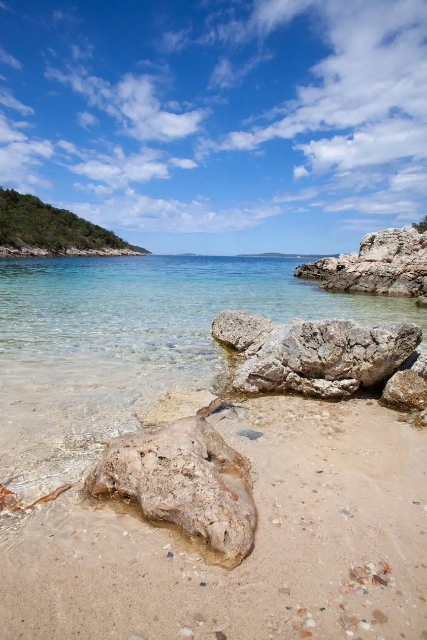 A scenic view of a clear, turquoise sea gently lapping against rocky shores and a sandy beach, under a bright blue sky with scattered clouds.