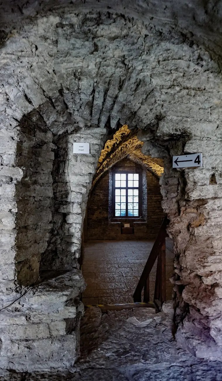 An arched stone doorway leading into a dimly lit room with a wooden staircase. The walls are rough-hewn stones, and a window is visible in the background with light filtering through.