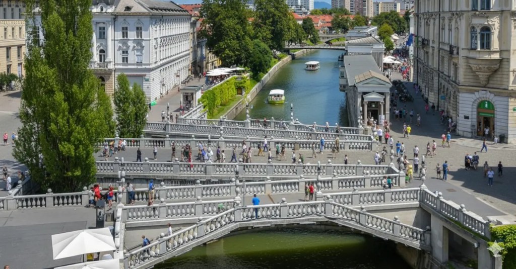 A panoramic view of Ljubljana, Slovenia, showcasing the vibrant cityscape with the Ljubljanica River bordered by lush greenery and historic buildings. The Triple Bridge is visible, bustling with people enjoying the sunny day.