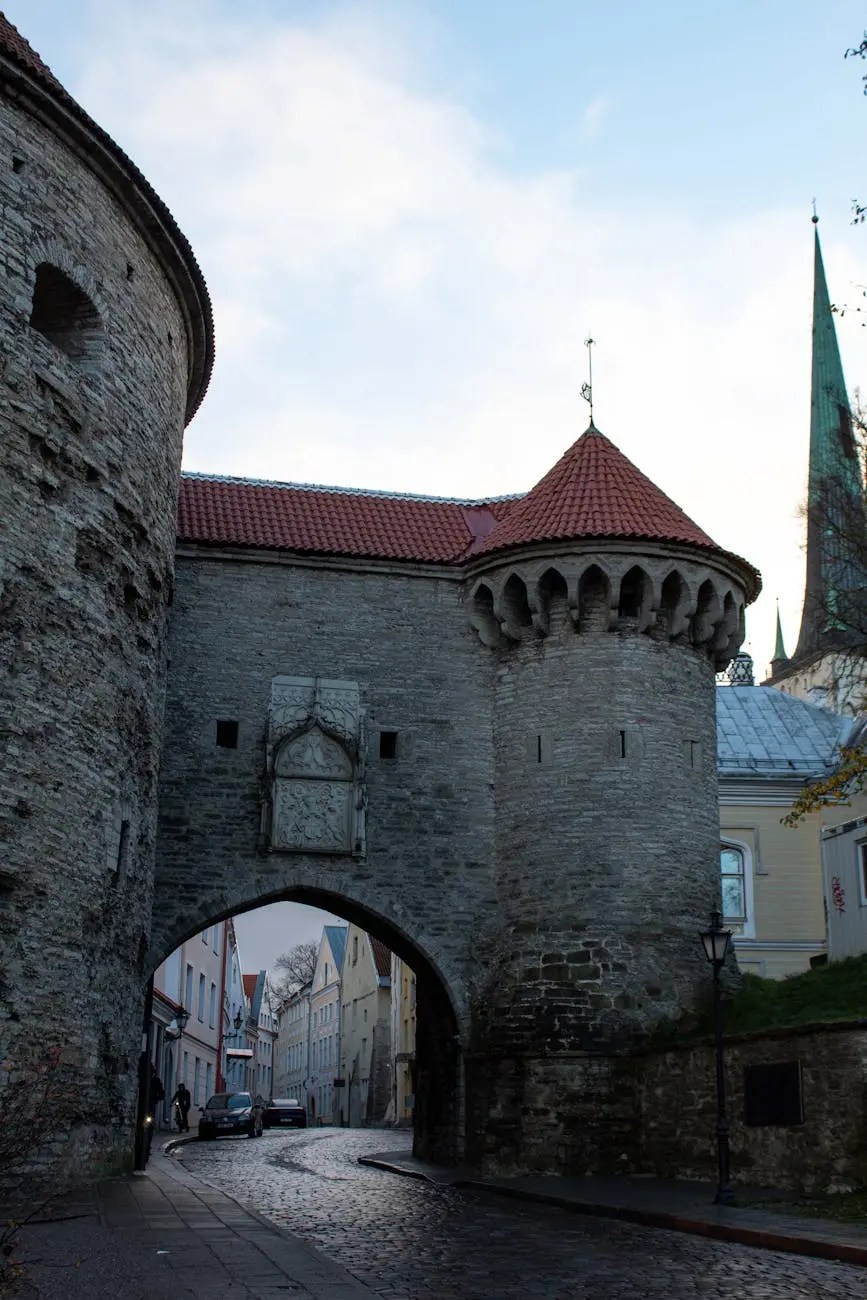 A view of a medieval stone archway with towers in Tallinn, Estonia, leading into a cobbled street lined with pastel-colored buildings.