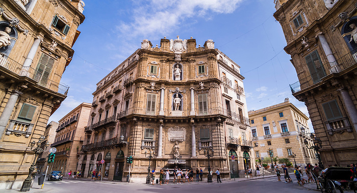 A historic building at the intersection of two streets in Palermo, featuring Baroque architecture and sculptures on the facade, with pedestrians and vehicles in the foreground against a bright blue sky.