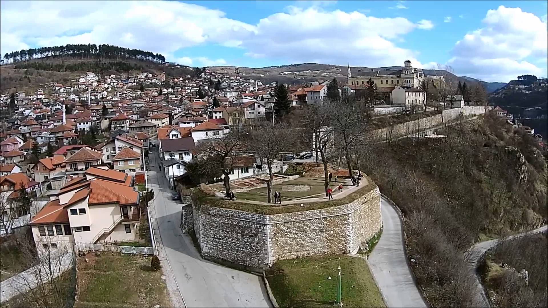 Aerial view of Sarajevo showcasing city rooftops with red-tiled roofs, green trees, and a distant mountainous landscape under a partly cloudy sky.