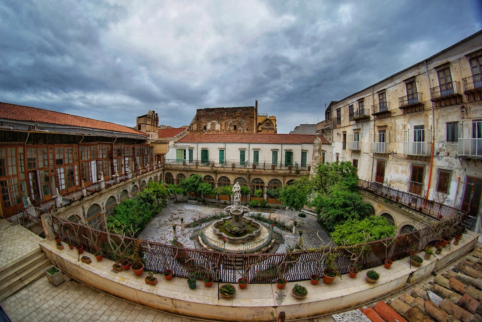 A view of a beautifully designed courtyard featuring a central fountain surrounded by greenery and historic buildings with balconies under a cloudy sky.
