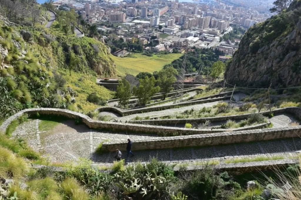 A pilgrim hiking the historic Acchianata stone path up Monte Pellegrino in Palermo, Sicily, with views of the Mediterranean Sea and surrounding hills.