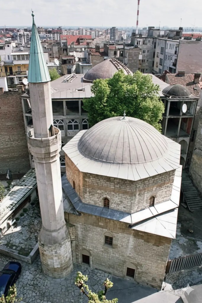 An aerial view of a historic mosque featuring a stone structure with a domed roof and a minaret, surrounded by modern buildings and greenery in Belgrade, Serbia.