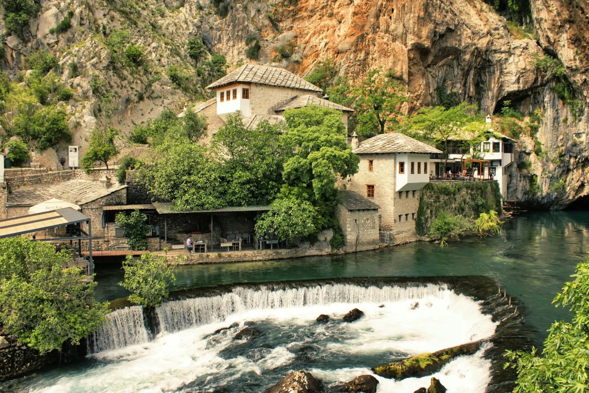 A scenic view of the Blagaj Tekija, a historic Dervish monastery nestled against a rocky cliff, with green trees and blue waters flowing beneath it.