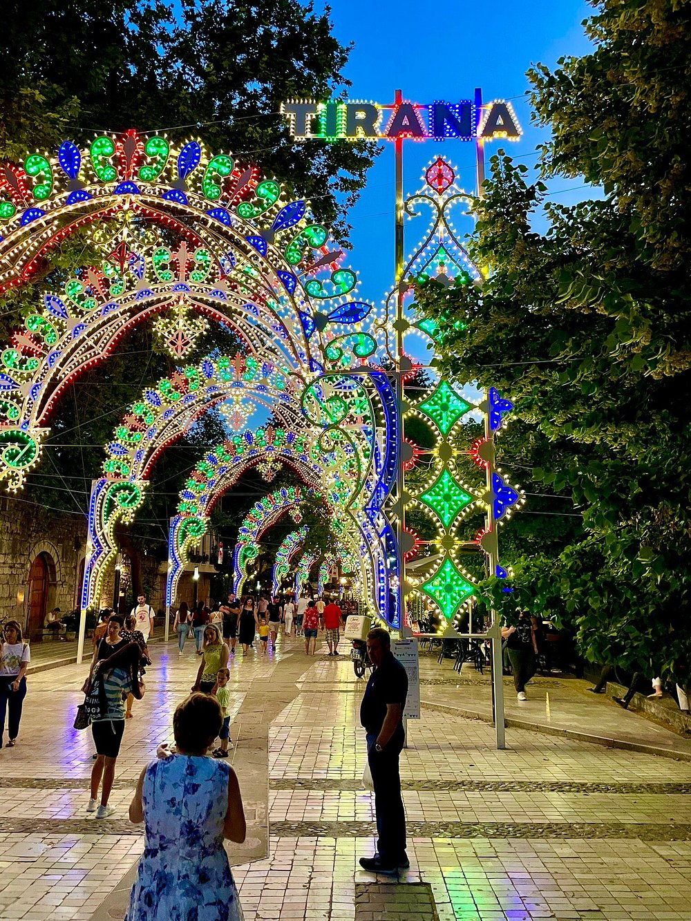 A vibrant street scene in Tirana, Albania, adorned with colorful lights and decorations. People stroll along a well-lit path, enjoying the lively atmosphere during the evening.