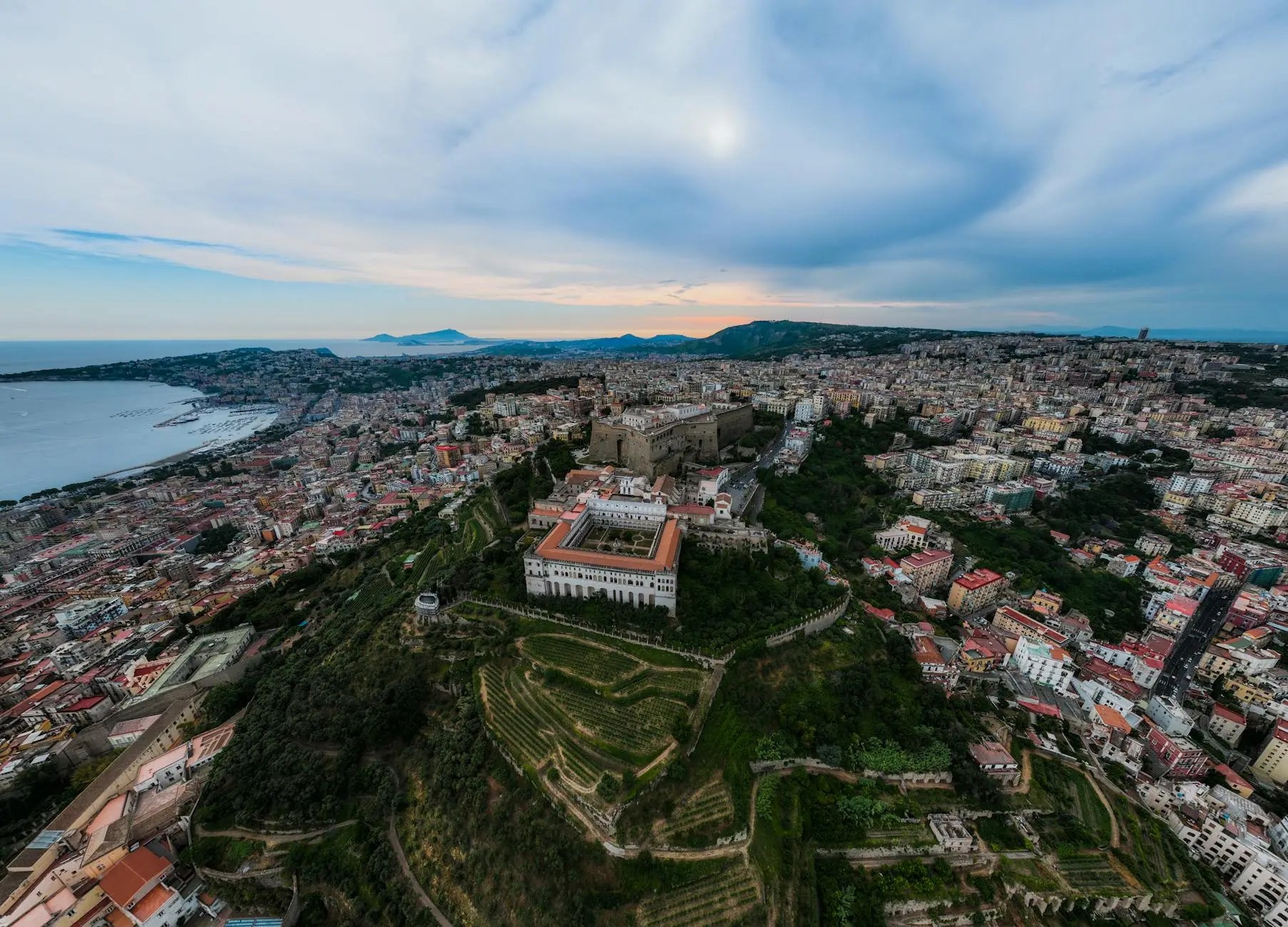 Aerial view of Naples with the coastline and hills, showcasing a mix of urban and natural scenery, including buildings, greenery, and the distant silhouette of mountains.