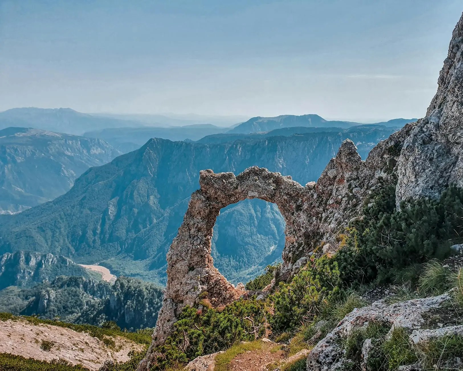 A rocky arch formation on a mountain with expansive valleys and hills in the background under a clear blue sky.