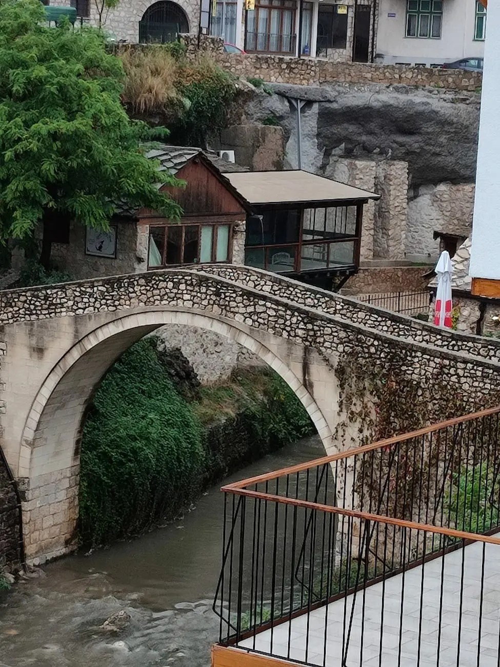 A stone arch bridge crosses a small river, surrounded by lush greenery and traditional buildings in Mostar, Bosnia and Herzegovina.