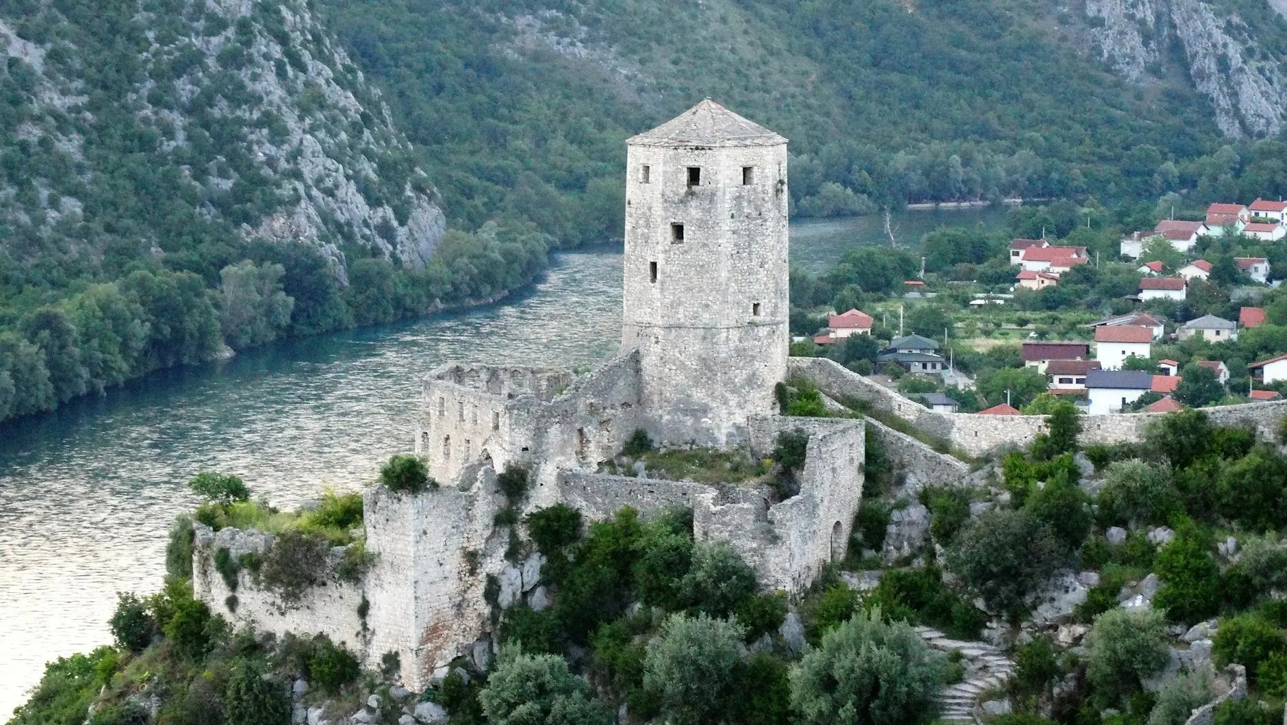 Aerial view of a historic fortress on a rocky hill overlooking a river, with lush greenery and houses in the background.