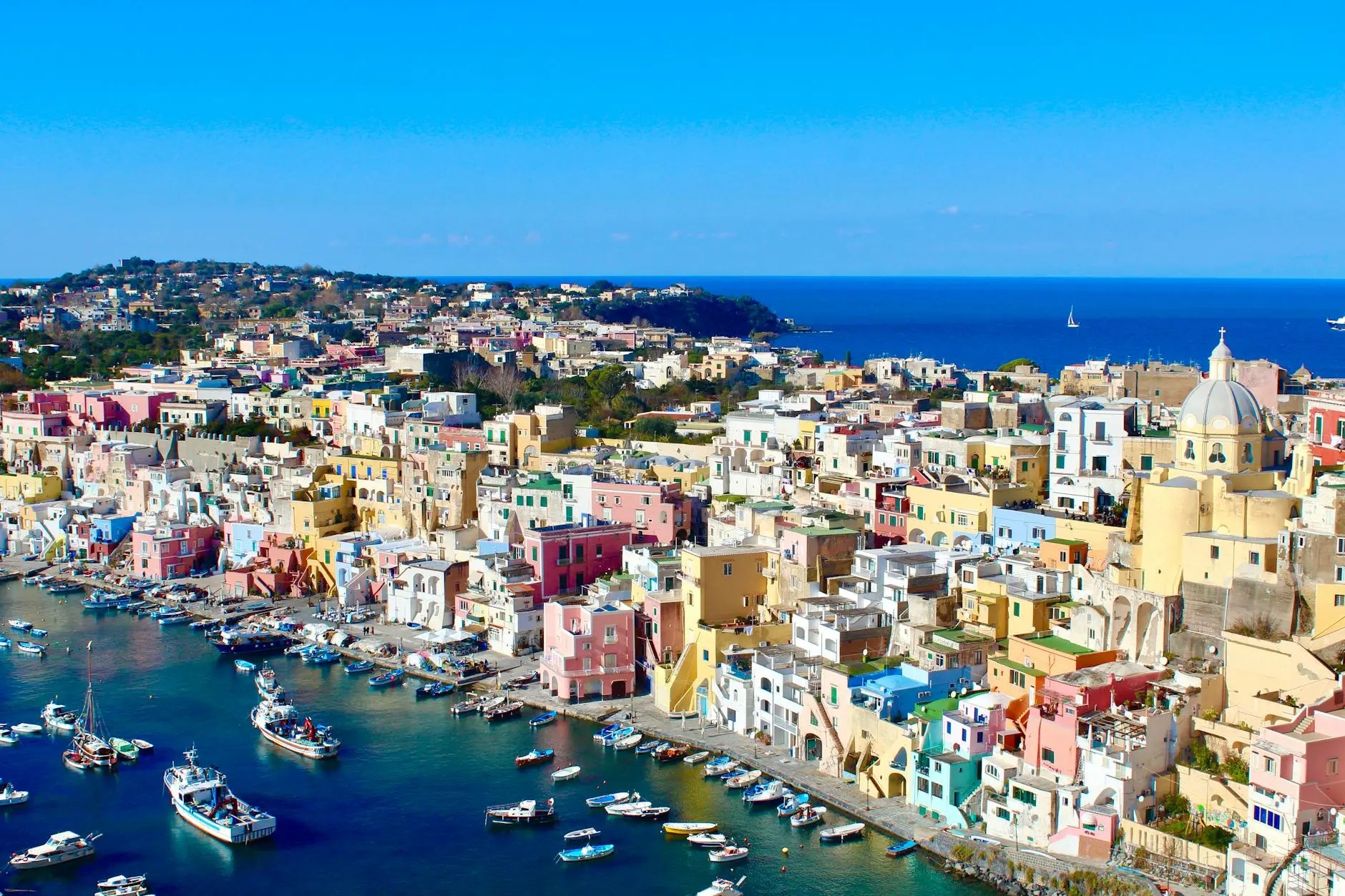 A scenic view of Procida, Italy, showcasing colorful buildings along the waterfront with boats docked in a clear blue bay and the Mediterranean Sea in the background.