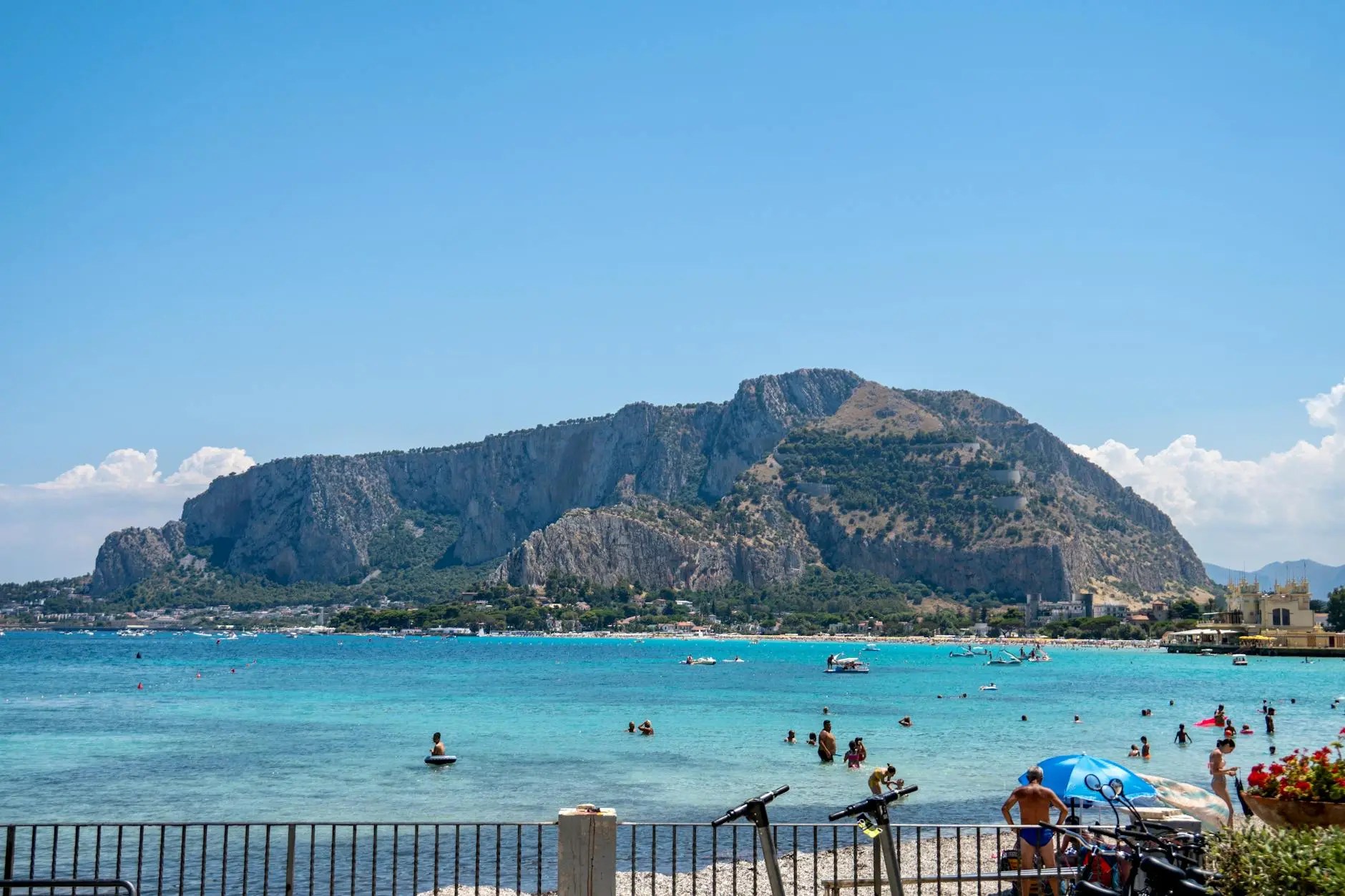 A panoramic view of Mondello Beach in Sicily, showing its iconic crescent of white sand and vibrant turquoise Mediterranean waters, backed by historic Liberty-style villas and green hills.