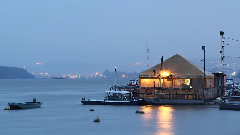 View of a river barge and a restaurant at dusk by the river, with city lights in the background.