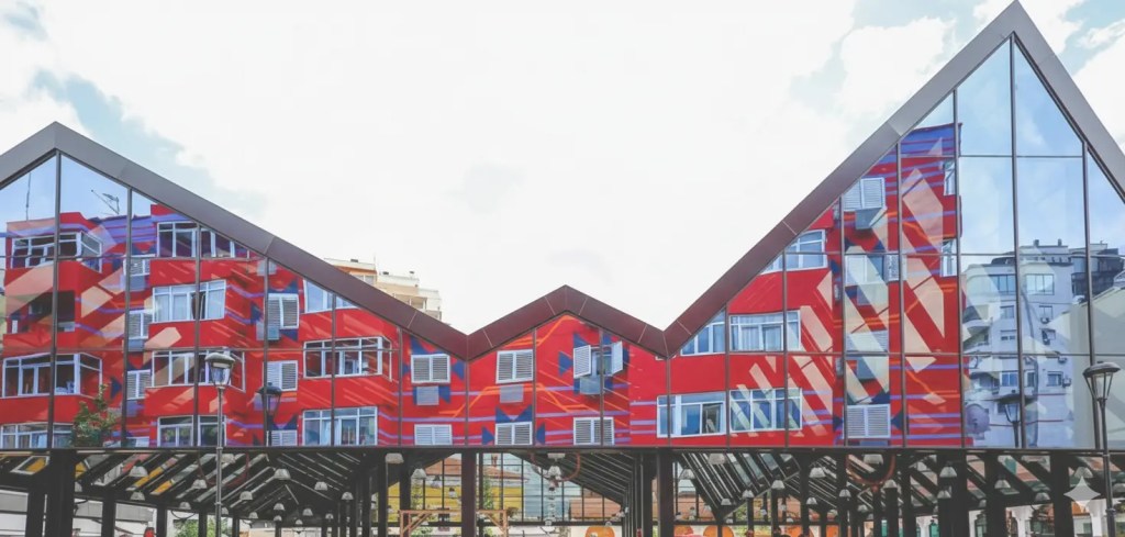 A colorful modern building with a distinctive peaked roof and reflective glass, showcasing vibrant red patterns, situated in Tirana, Albania.
