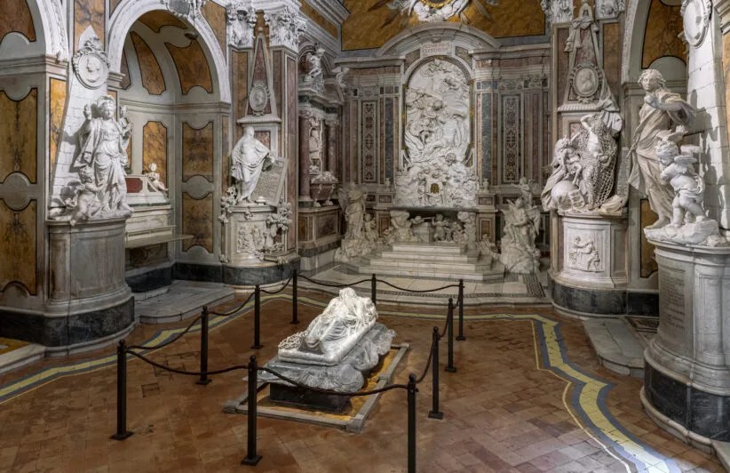 Interior view of the Cappella Sansevero in Naples, showcasing intricate marble sculptures and ornate decorations, with a prominent statue of the Veiled Christ.