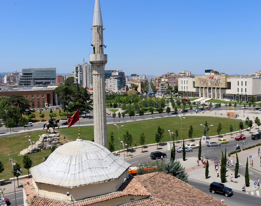 A panoramic view of Tirana, Albania, showcasing the iconic clock tower and the National Historical Museum in the distance, with a vibrant cityscape featuring modern buildings and green spaces.
