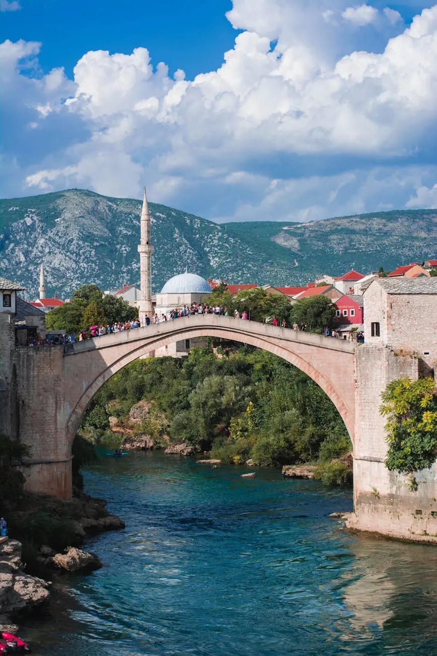 The historic Stari Most bridge in Mostar, Bosnia and Herzegovina, spans the Neretva River with a backdrop of mountains and traditional architecture under a bright blue sky.