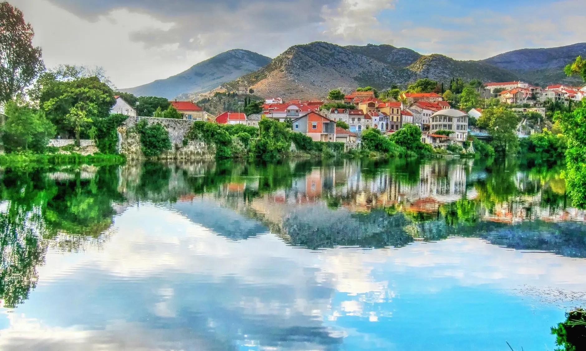 "A scenic aerial view of Trebinje, the southernmost city in Bosnia and Herzegovina, nestled along the calm, reflective waters of the Trebišnjica River in a green valley."