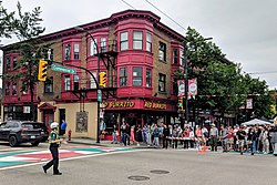A busy street scene with a red-brick building on the corner and a crowd of people gathered nearby. A police officer directs traffic while festival banners and tents are visible.