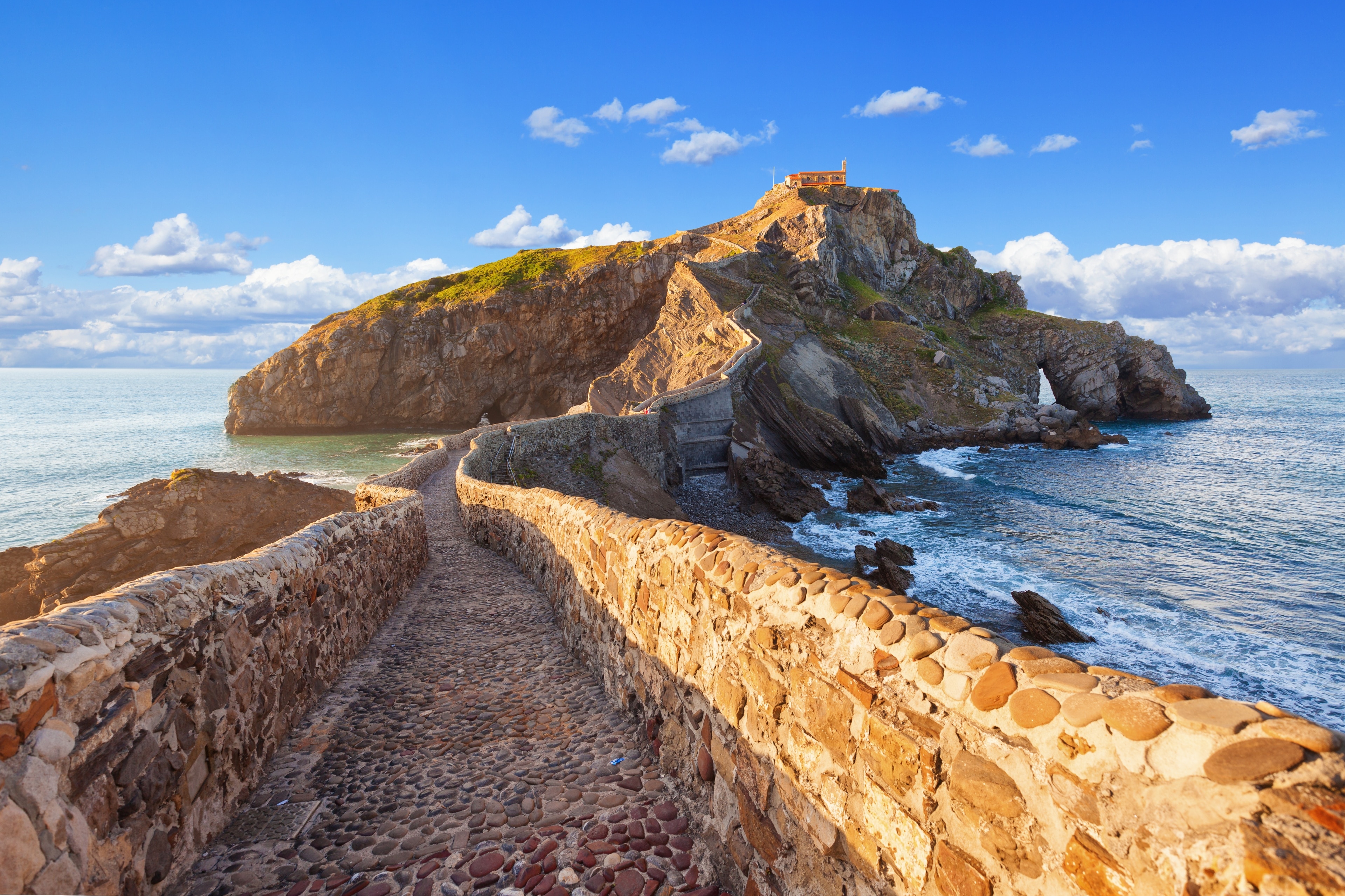 A scenic view of a rocky island connected to the shore by a stone path, under a clear blue sky with clouds.
