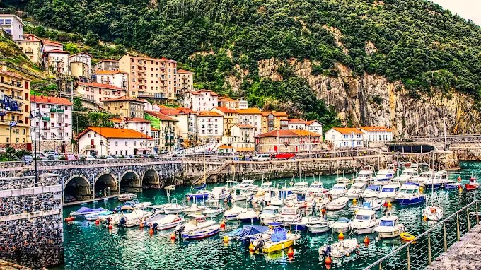 A picturesque coastal view featuring a marina filled with various boats, surrounded by colorful buildings and lush green hills.