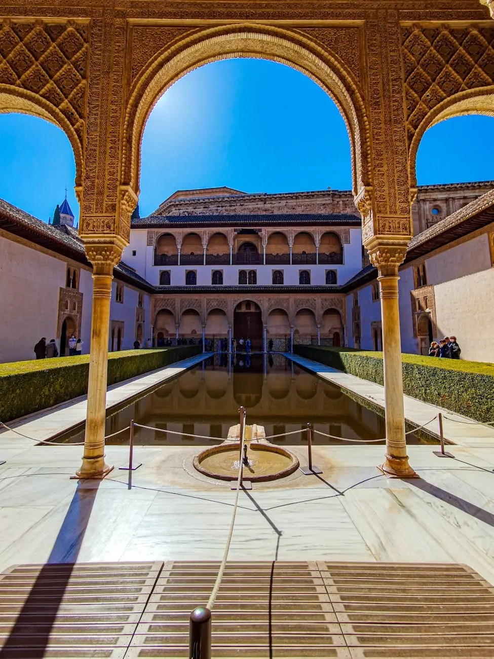View of the Courtyard of the Lions in the Alhambra, featuring intricate arches, a central fountain, and lush greenery under a clear blue sky.