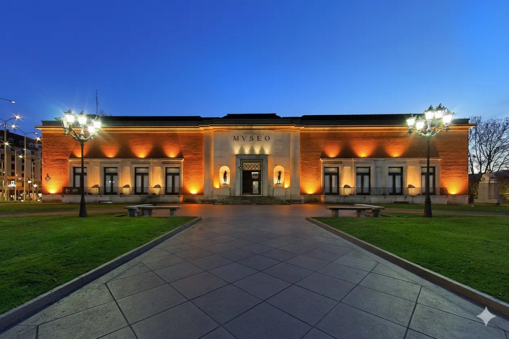 Exterior view of a museum building illuminated at dusk, featuring orange lighting highlights and a grassy area in front.