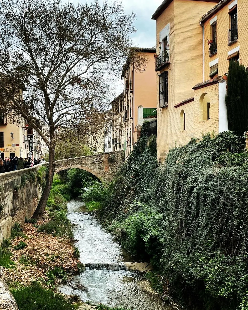 A picturesque view of a narrow stream running alongside a stone bridge, surrounded by lush green vegetation and charming buildings in Granada, Spain.