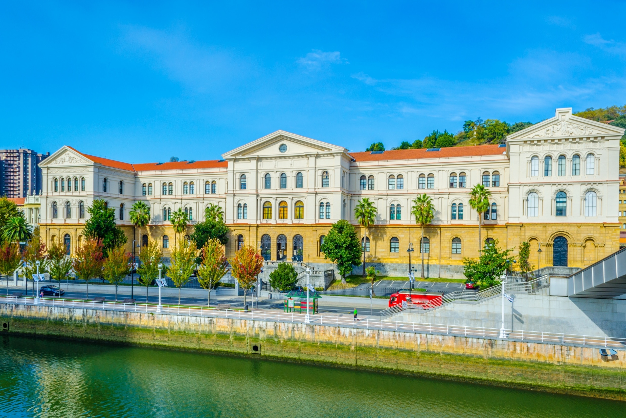 View of a historic building with a neoclassical facade, surrounded by palm trees and greenery, next to a river under a clear blue sky.