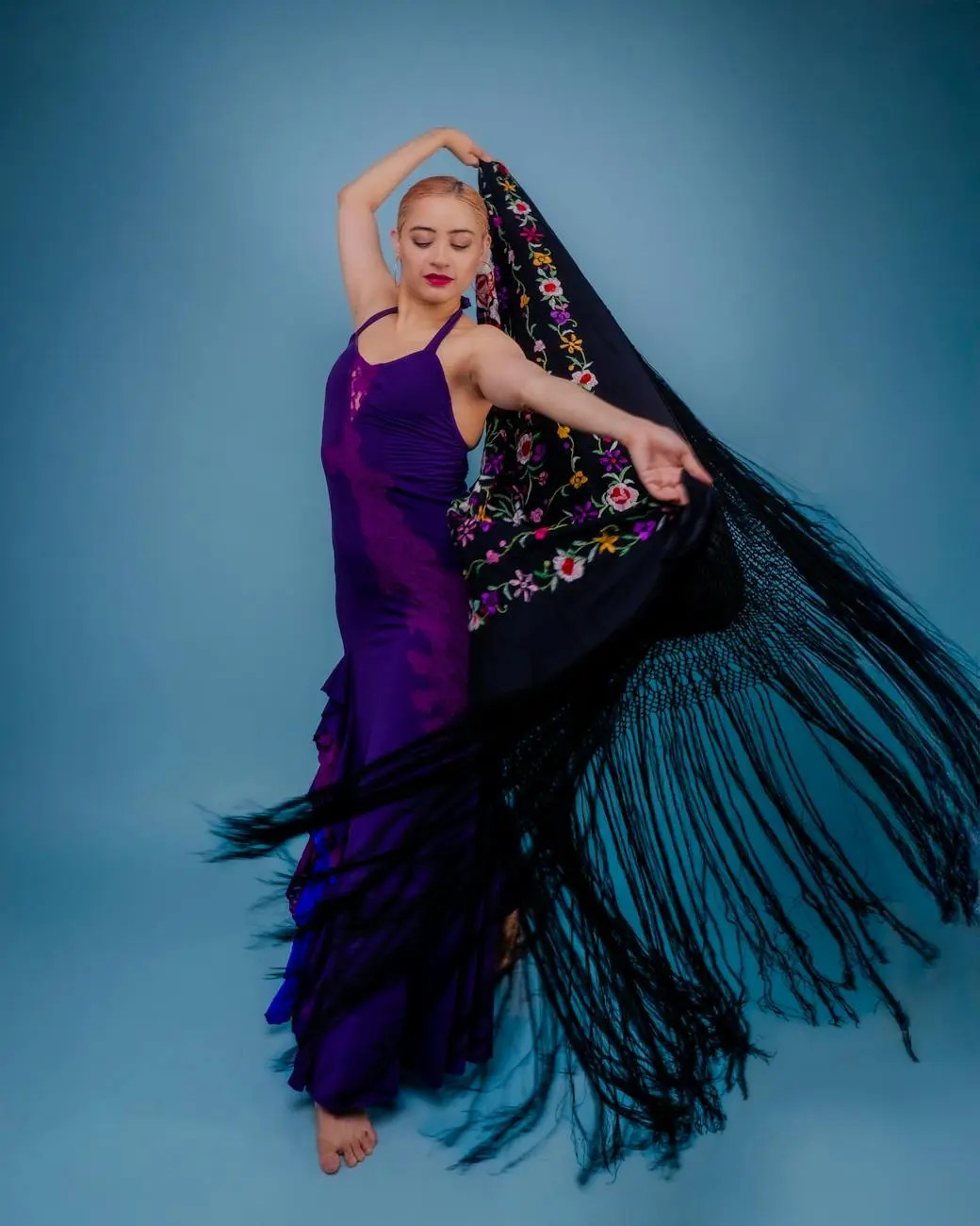 A dancer in a purple dress gracefully twirls with a flowing black shawl against a solid blue background.