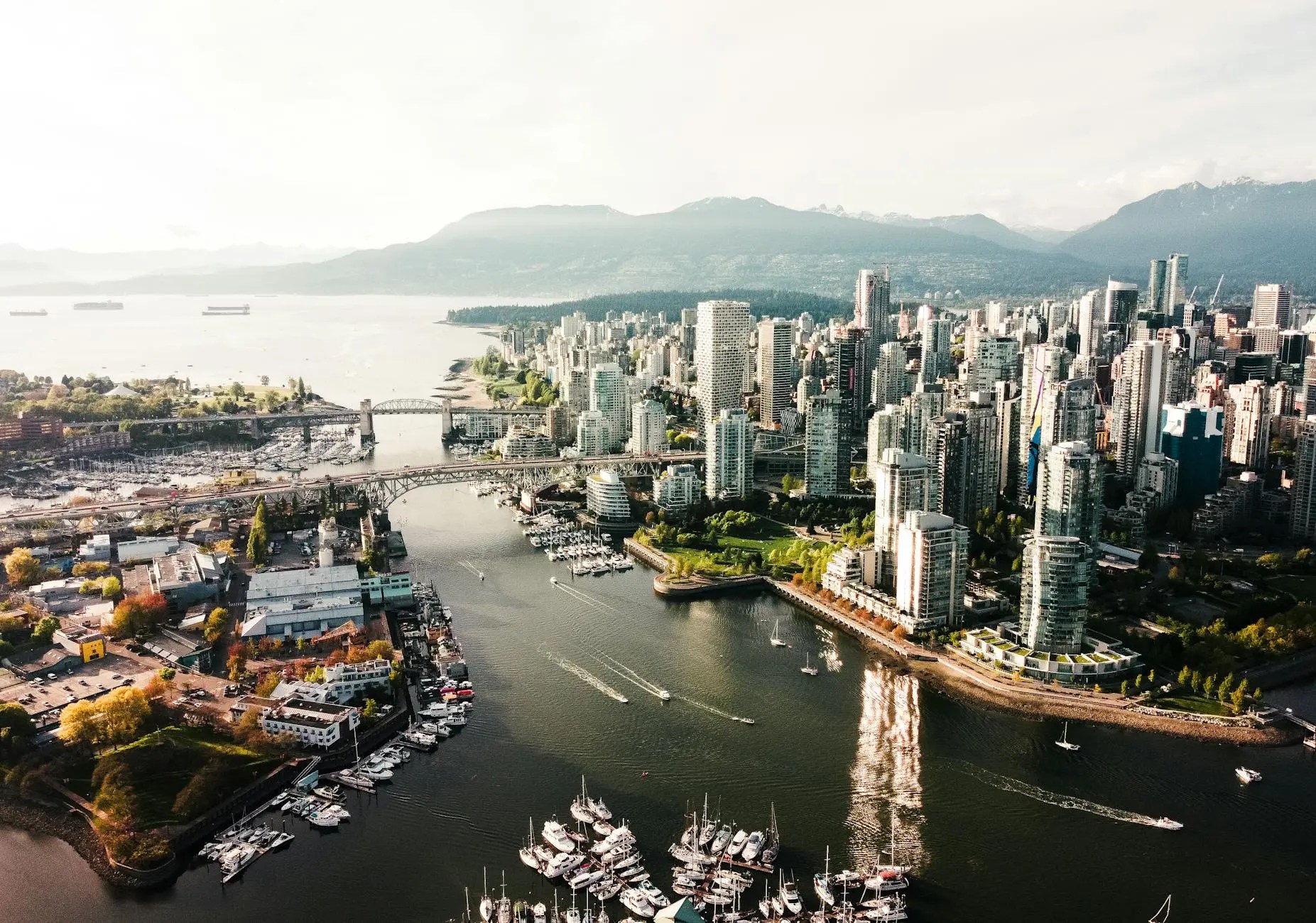 Aerial view of Vancouver city showcasing modern skyscrapers, a marina filled with boats, a bridge crossing the water, and mountainous terrain in the background.
