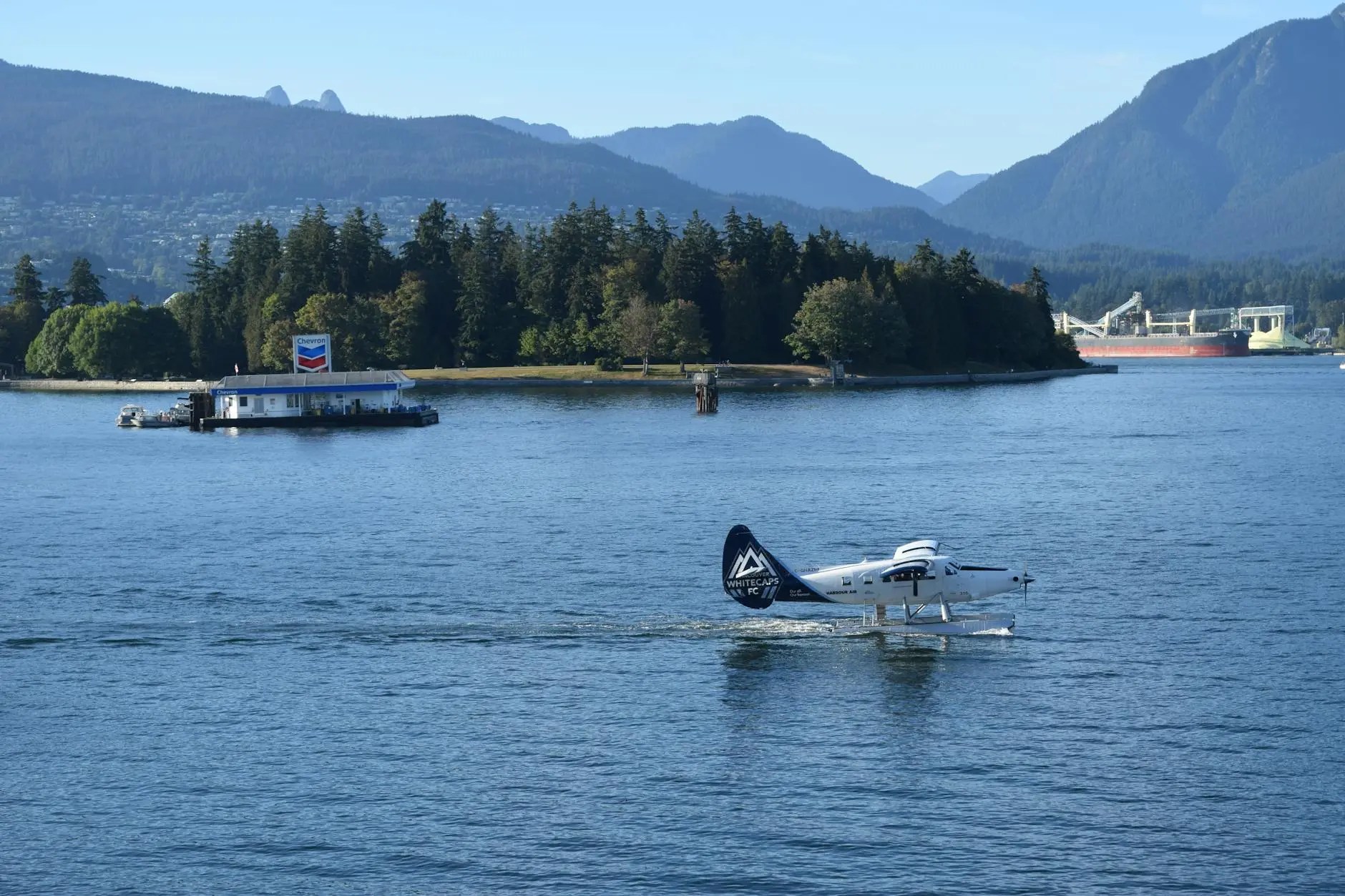 A seaplane cruising over water with a Chevron fuel station in the background, surrounded by lush greenery and mountains under a clear blue sky.