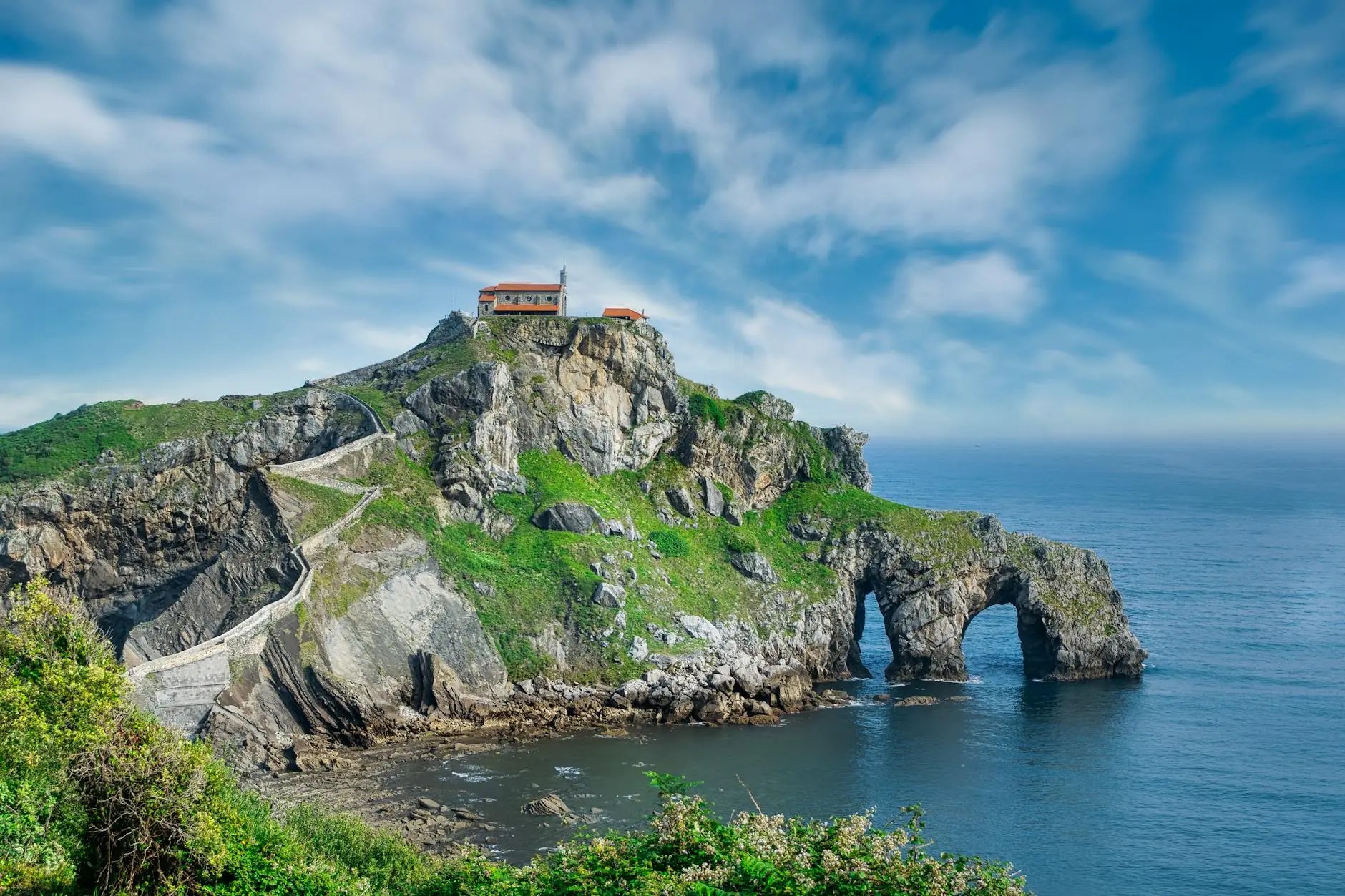 Scenic view of a rocky cliff with a small house on top, surrounded by greenery and overlooking the ocean, under a partly cloudy sky.