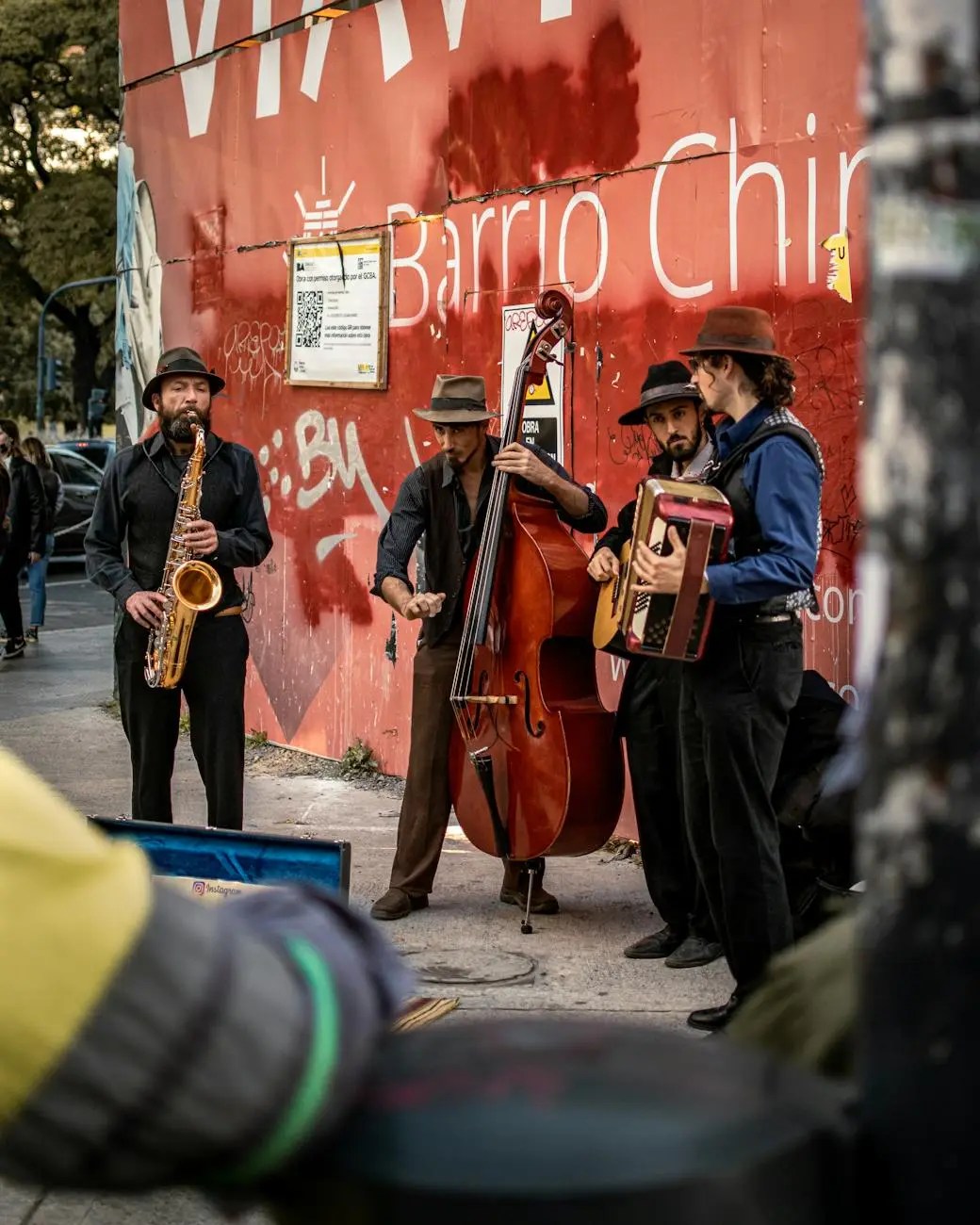A group of four musicians performing on the street, featuring a saxophonist, a double bassist, an accordionist, and a guitarist, in front of a red wall with graffiti.